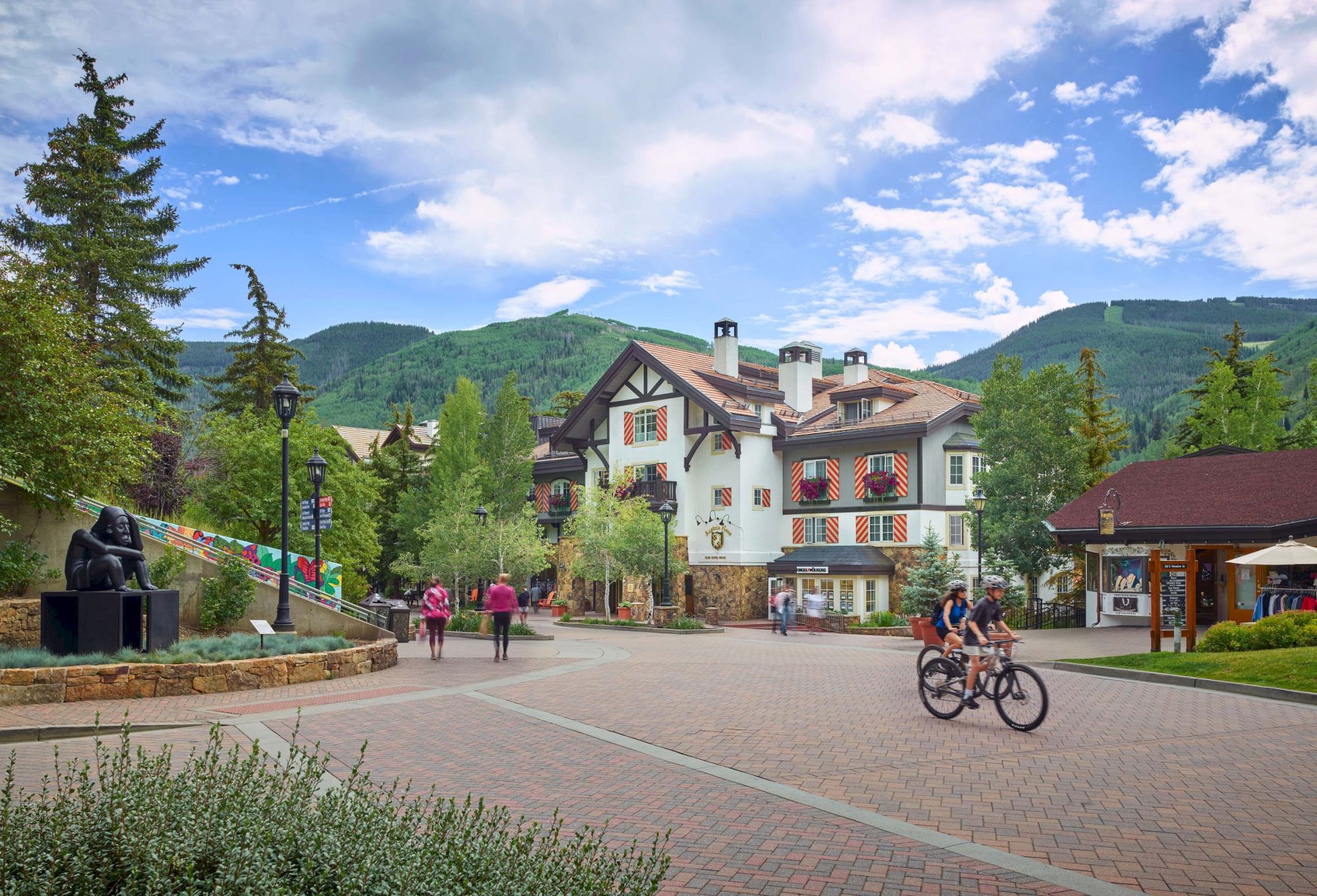 A picturesque alpine village square with a white chalet, cyclists, people strolling, green hills, and blue skies, sunny and peaceful.