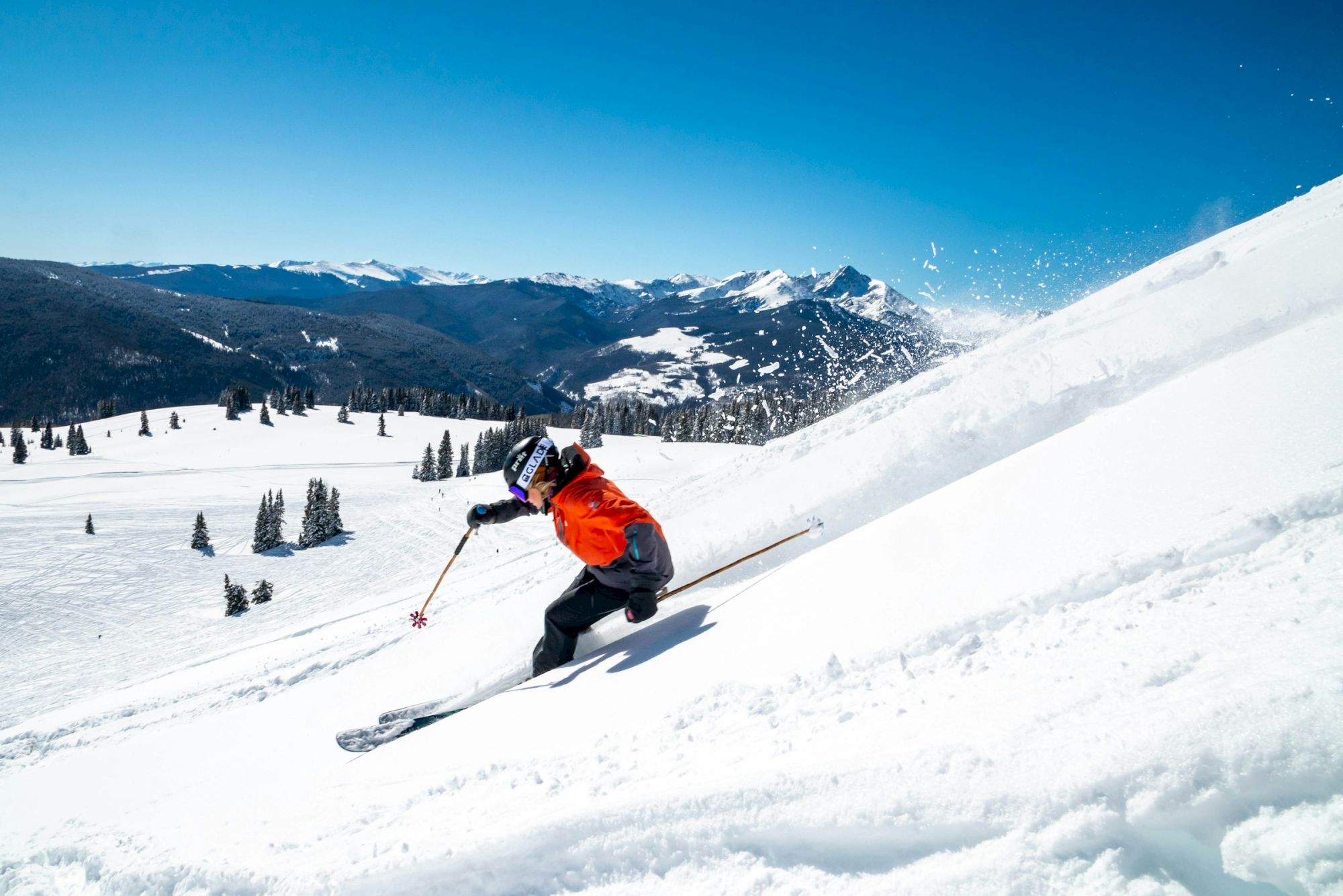 A skier in an orange jacket carving down a snowy slope, mountains in the background under a clear blue sky.