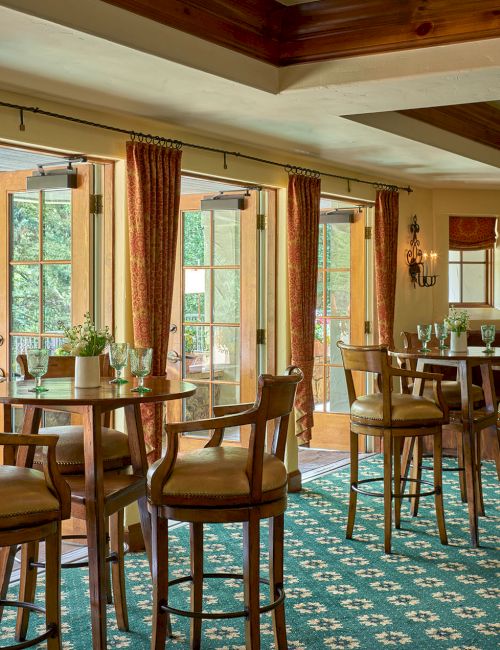 The image shows a cozy dining area with wooden tables and chairs, patterned carpet, large windows, and natural light.