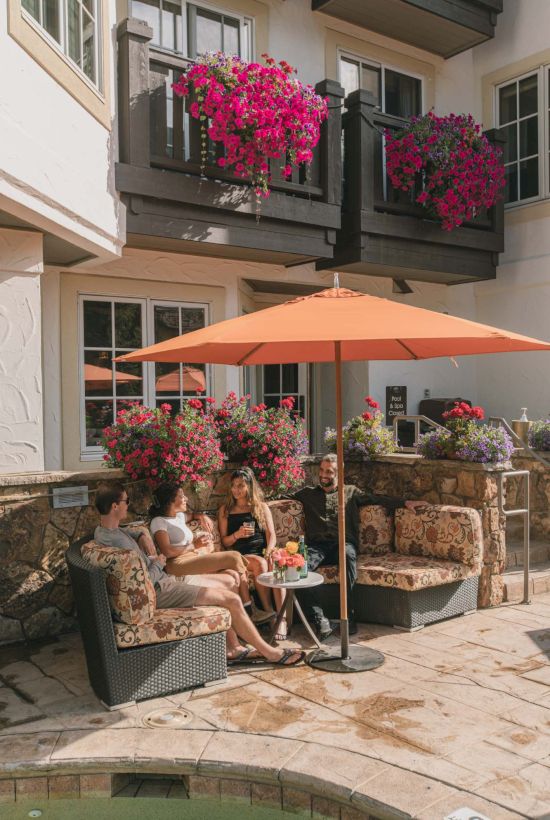 People relax on a patio with a central umbrella, seating, and vibrant hanging plants on the building&rsquo;s balconies, sunny and inviting.