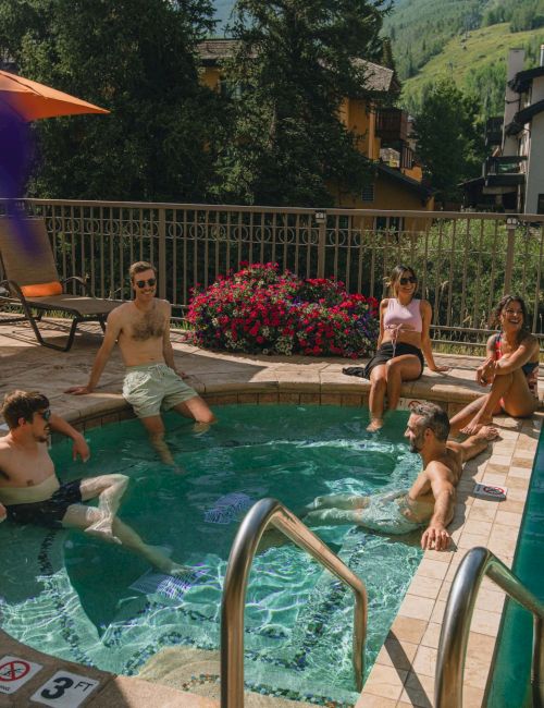 Several people relax in and around a pool on a sunny day, some seated on the edge, others in the water, with a hillside resort and orange umbrella nearby.