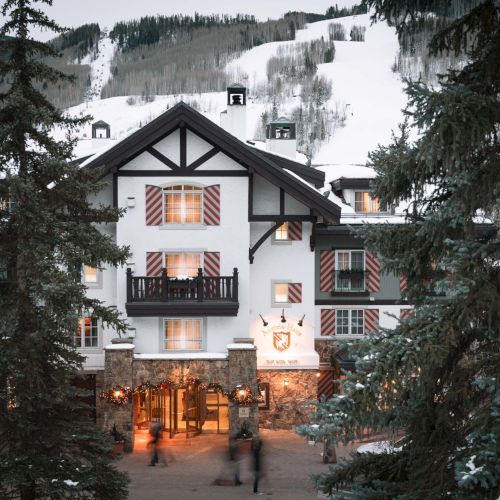 A quaint two-story cottage with a steep roof, warm lights glow from windows, surrounded by tall trees and autumn foliage.