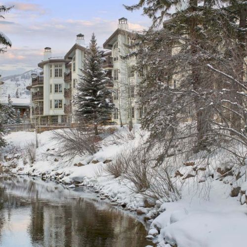 A snowy village scene with a stone bridge and houses blanketed in fresh snow, evergreen trees dusted white, and a quiet winter morning.