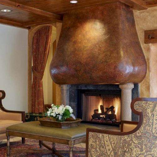 A cozy living room with a large copper fireplace, two patterned armchairs, a wooden coffee table, a side desk, and a warm, rustic d&eacute;cor.