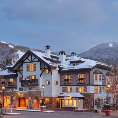 A snowy alpine village at dusk with warmly lit chalets, mountains in the background, and a calm, festive feel.