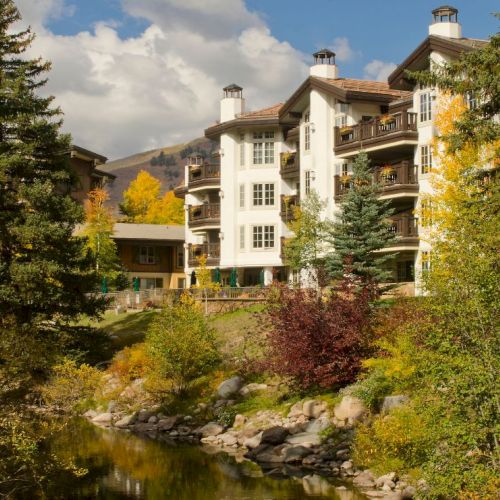 A charming white and beige hillside hotel or apartment building nestled among autumn trees and greenery. Top it at 140 characters, always ending the sentence.