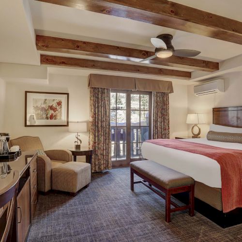 A hotel room with two queen beds, rustic wood beams, a sitting area, desk, TV, balcony doors, and warm lighting.