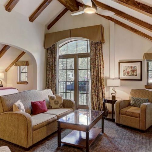 A cozy living area in a rustic suite with a fireplace, sofa, chairs, coffee table, a TV, and a bedroom visible through an arched opening.