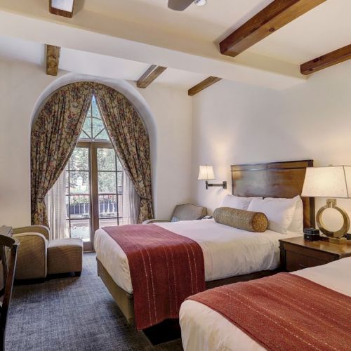 A hotel room with two queen beds, matching headboards, lamps, a seating area by a window with arched drapes, and neutral decor with red bed runners.