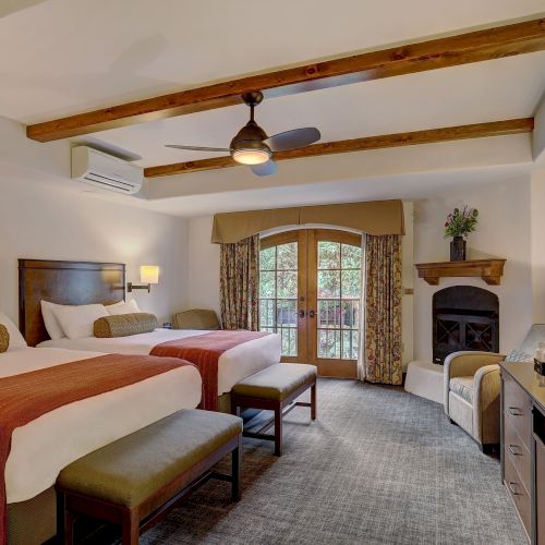 A spacious hotel room with two queen beds, a warm rustic decor, a fireplace, vaulted ceiling with wooden beams, and a balcony view.