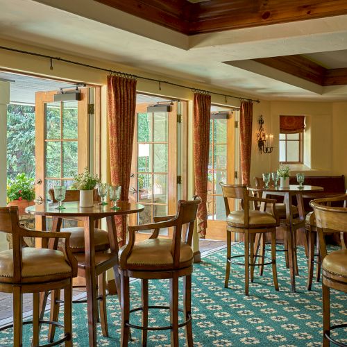 A cozy restaurant with wooden chairs and round tables, large windows with red curtains, and a green patterned carpet, creating a warm dining area.