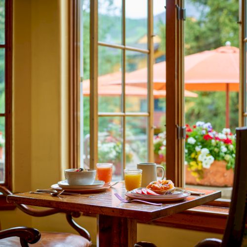 Cozy breakfast nook by a sunny window; a wooden table set with coffee, orange juice, and pastries, overlooking a garden with orange umbrellas.