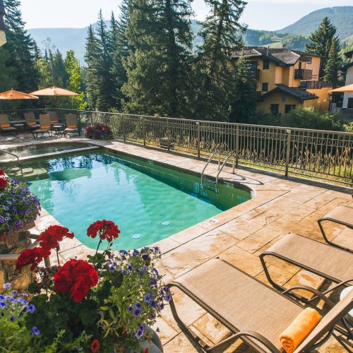 A sunny pool deck with lounge chairs, orange umbrellas, potted flowers, and a mountain-view courtyard surrounding a rectangular swimming pool.