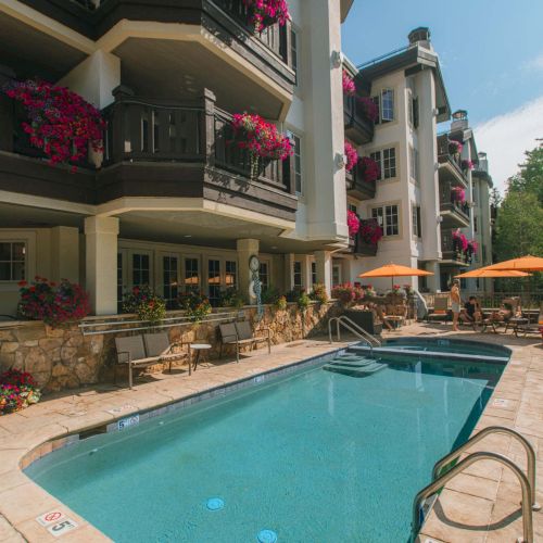 A resort-style swimming pool with clear blue water, bordered by lounge chairs, balconies with flower boxes, and orange umbrellas in a sunny courtyard.