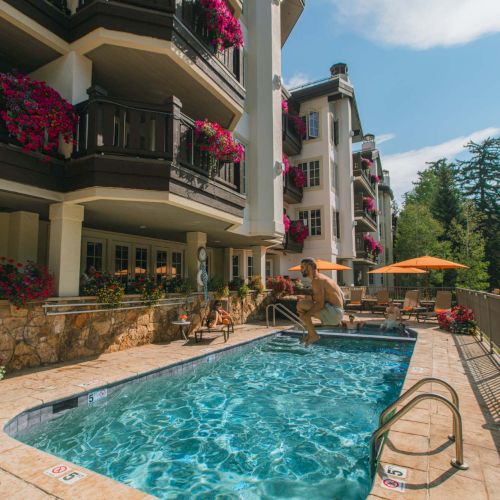 A multi-story apartment building with balconies adorned by pink flowers overlooks a rectangular pool with clear blue water, lounge chairs, and orange umbrellas.