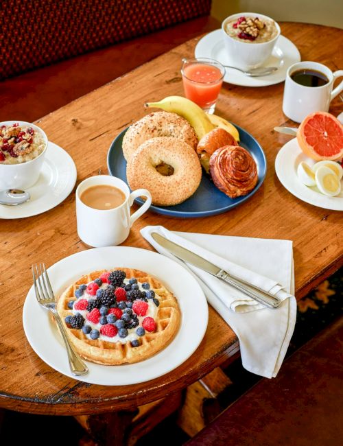 A cozy breakfast spread: waffles with berries, bread, croissant, fruit plates, coffee, juice, tea, and orange slices on a wooden table.