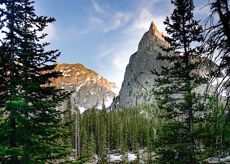 Snowy mountain peak rises above a pine forest, reflected in a tranquil lake at dusk.