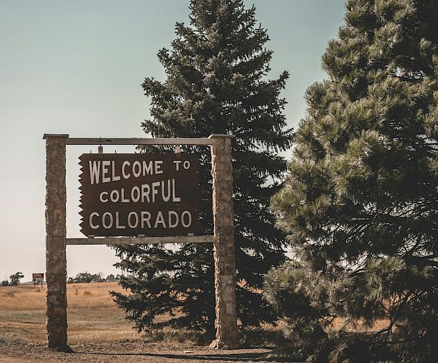 A weathered &ldquo;Welcome to Colorful Colorado&rdquo; sign stands between two pine trees on a dry, open field.