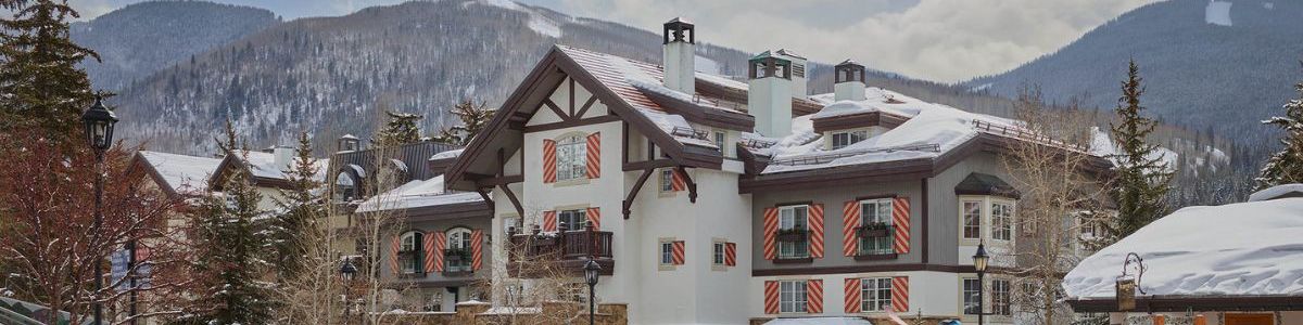 A snowy mountain village with chalet-style buildings, pine trees, and people walking on a sunny winter day.