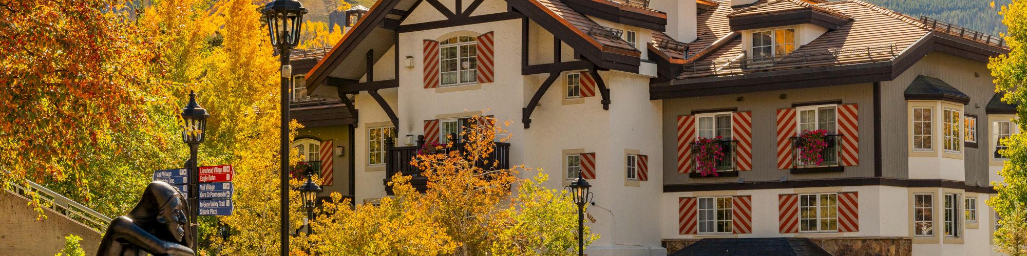 A picturesque alpine village with a charming white timbered inn, autumn trees, a statue, and a parked car against yellow mountains.