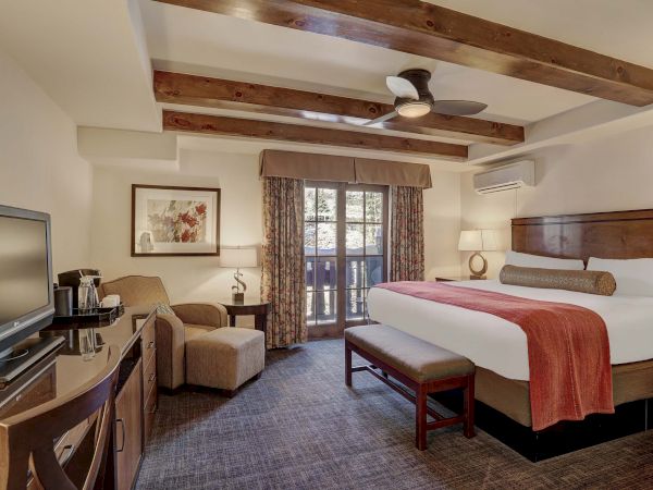 A cozy hotel room with a bed, TV, armchair, and a balcony. The decor features wooden beams and warm tones.