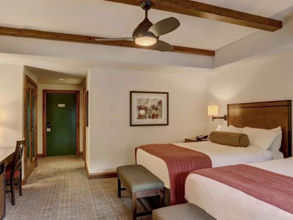 The image shows a hotel room with two double beds, a TV, desk, ceiling fan, and art on the wall, creating a cozy atmosphere.